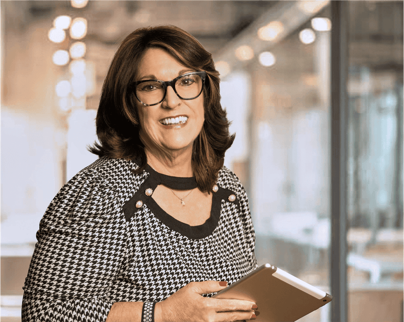 Smiling professional woman in houndstooth blouse and black-rimmed glasses holding a clipboard in a bright office setting