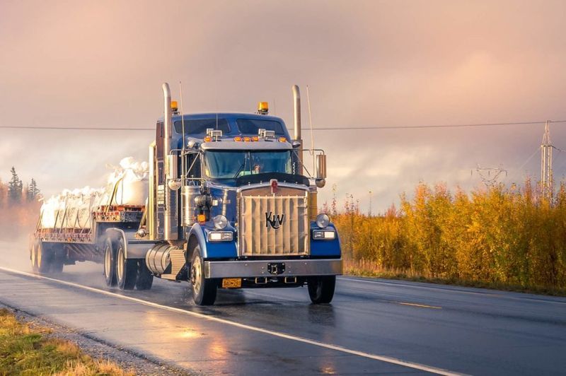 Blue semi-truck hauling flatbed trailer on rural highway at sunset