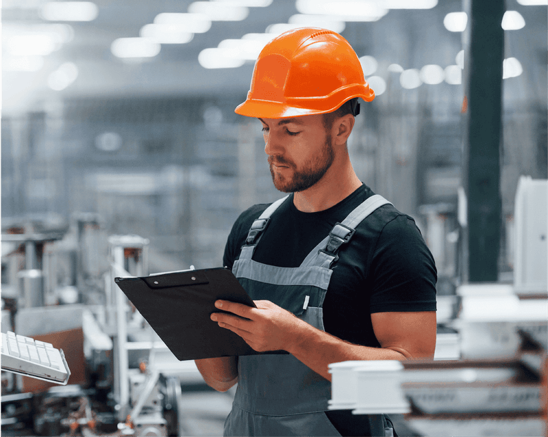 Worker in bright orange hard hat and safety vest reviews clipboard in modern industrial facility with machinery in background