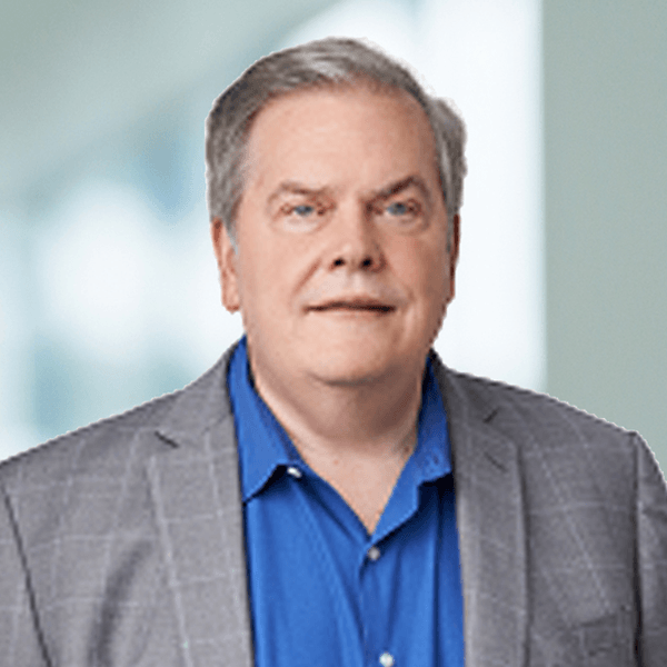 Middle-aged man with gray hair wearing a gray blazer and blue shirt, smiling confidently against a soft blue background