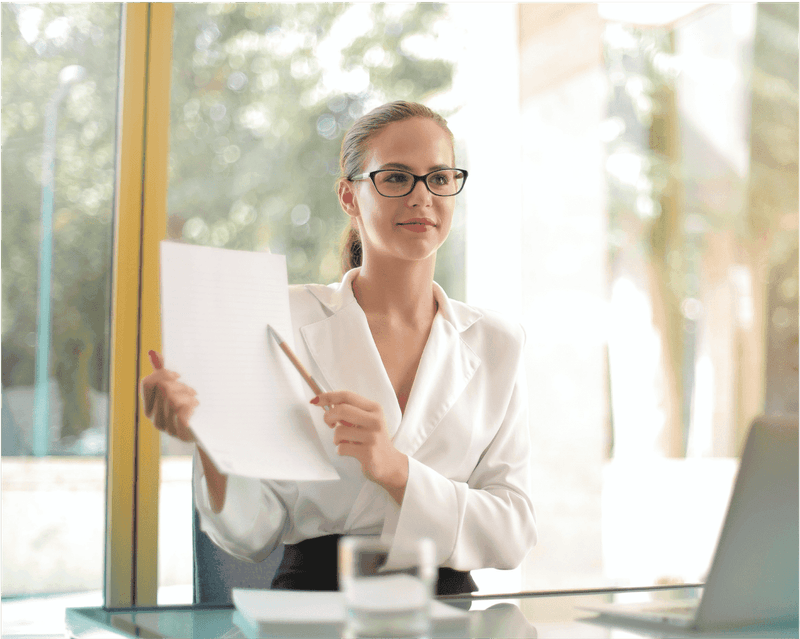 Professional in white blouse and glasses holds document while standing by window with laptop and glass of water