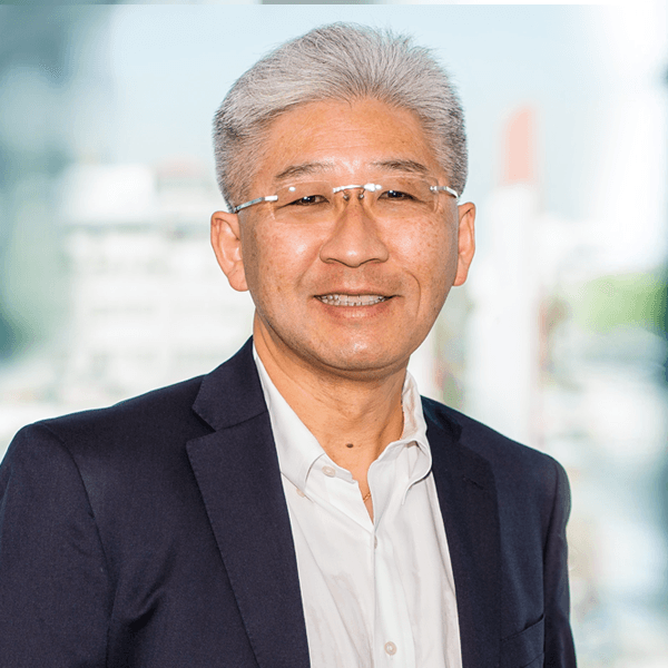 An older Asian professional with gray hair and rimless glasses smiles warmly while wearing a navy blazer and white shirt