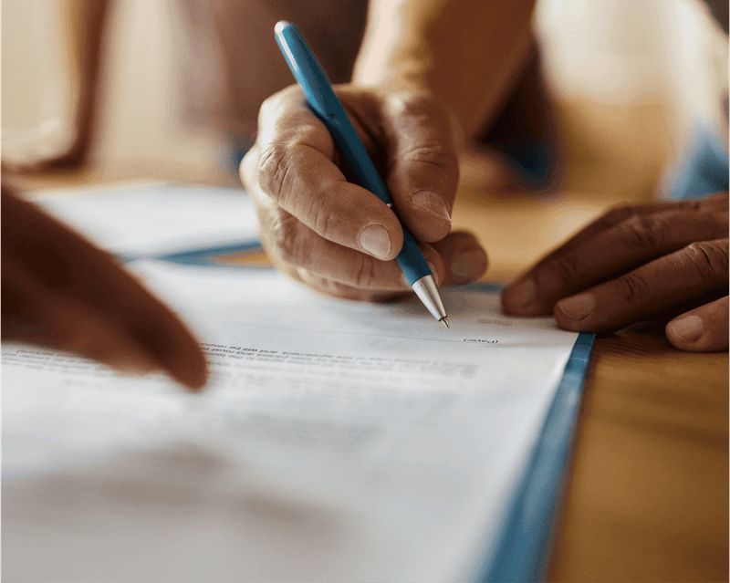 Hands with blue pen signing a white document featuring text visible on the paper and warm-toned background