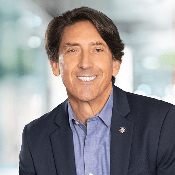 Smiling middle-aged professional in navy blazer and blue shirt featuring wavy dark hair, against a blurred office background