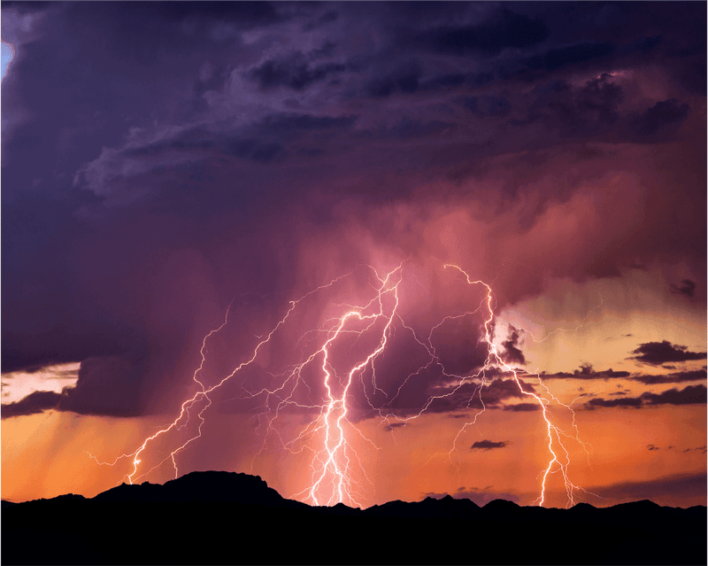 Lightning bolts strike dramatically from dark purple thunderclouds against an orange sunset sky over silhouetted