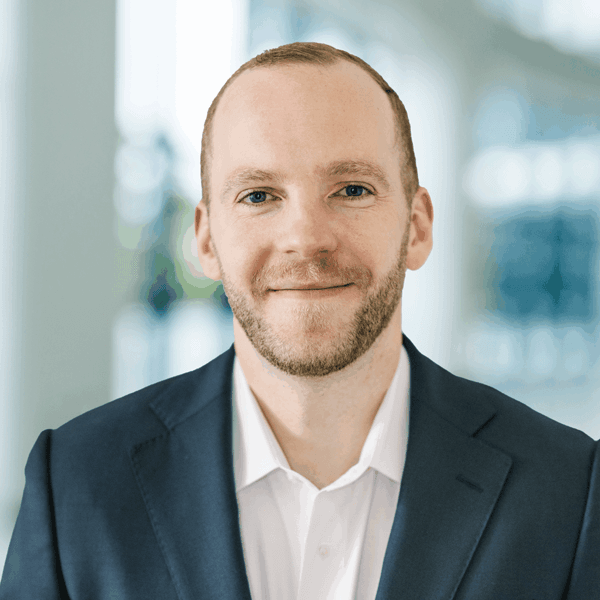 A smiling professional in a dark suit and white shirt featuring a blurry office background