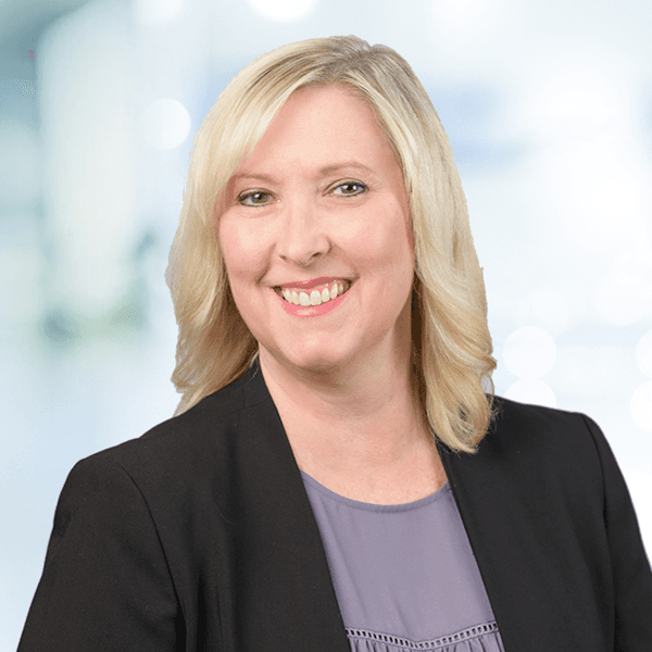 A blonde woman in a black blazer and gray top smiles warmly against a blurred office background